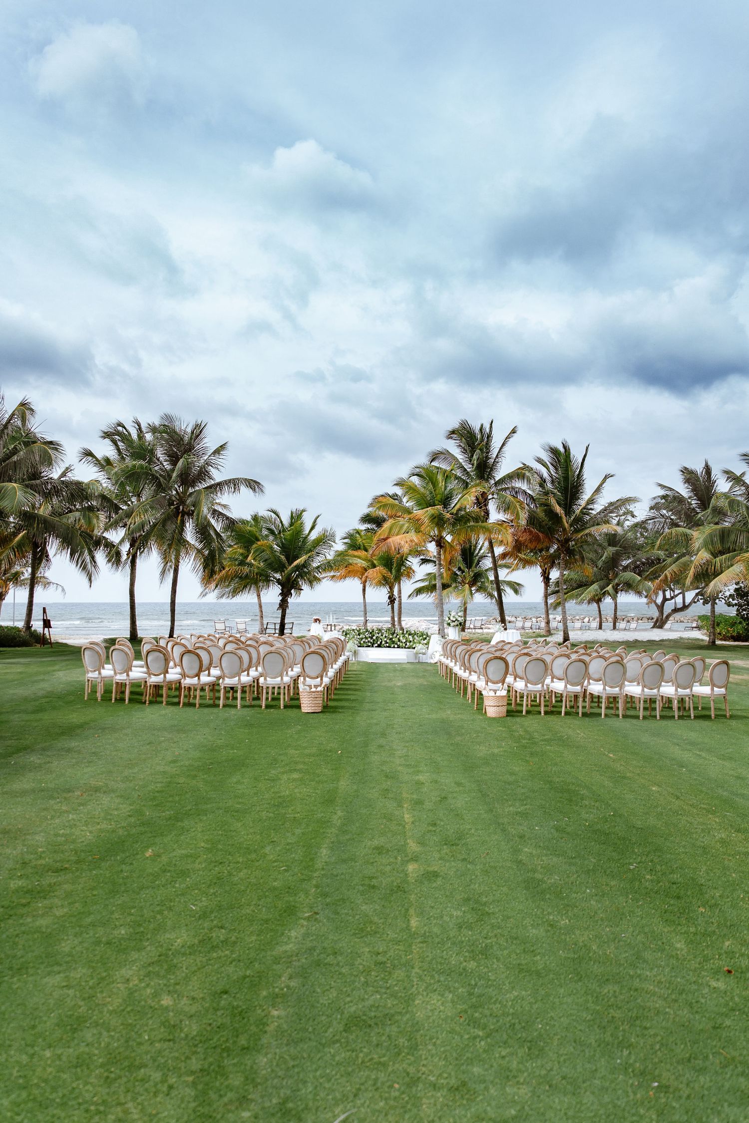 Outdoor beach wedding ceremony setup with elegant chairs, lush palm trees, and ocean views on a green lawn.