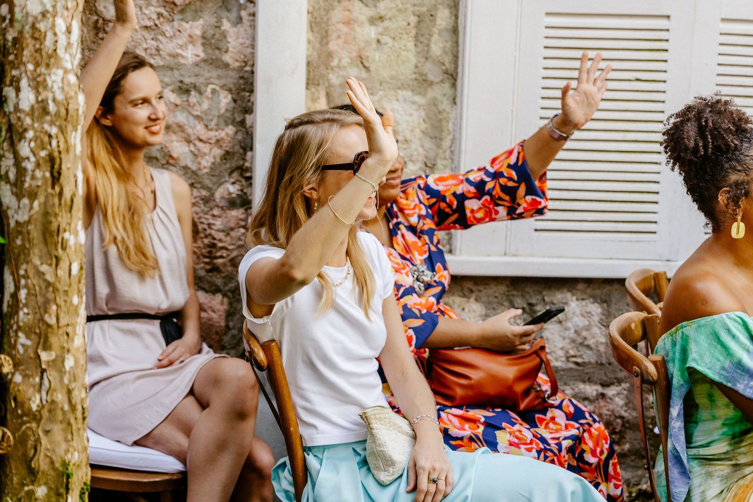 Four women socializing outdoors, laughing and waving, wearing colorful summer outfits near a rustic stone wall.