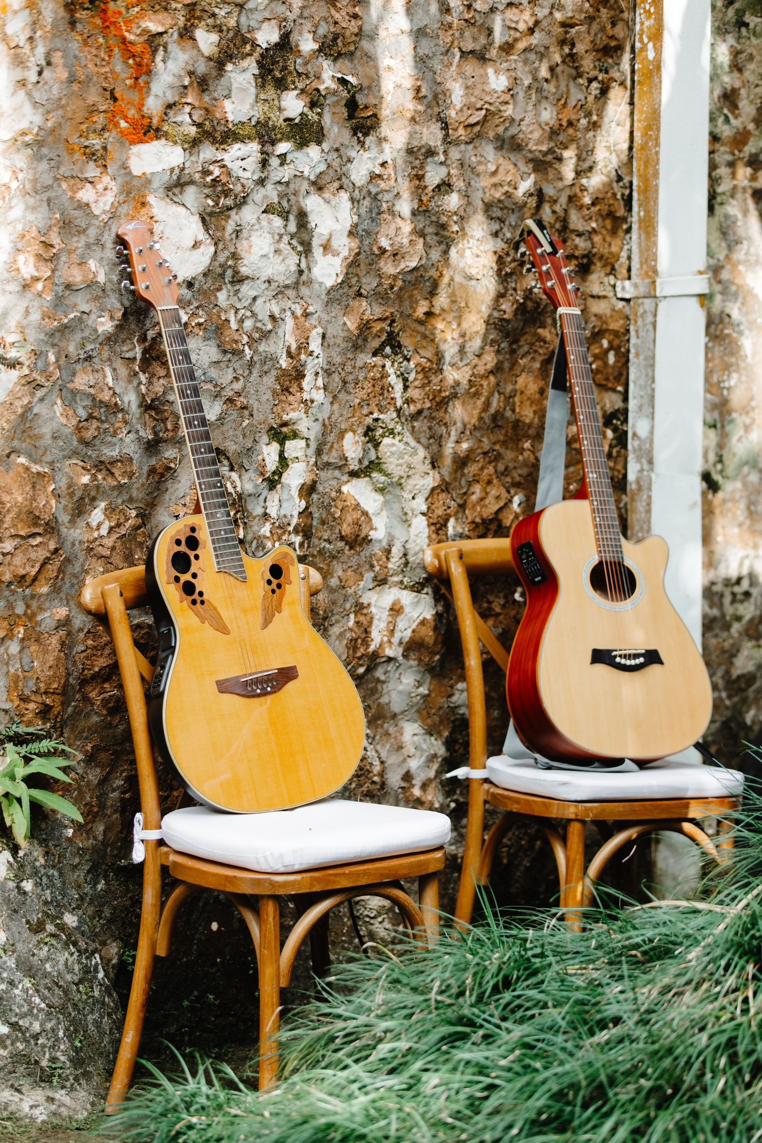 Two acoustic guitars resting on wooden cross-back chairs against a rustic stone wall outdoors.