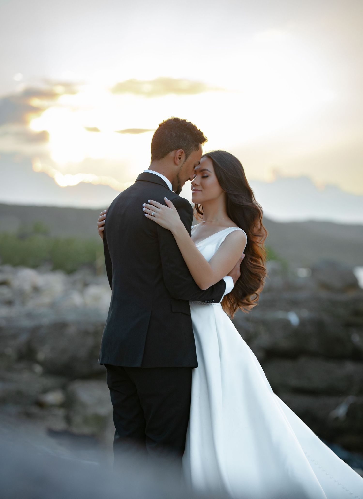 A bride and groom share a romantic embrace at sunset, with the bride in a flowing white gown and groom in a black tuxedo.