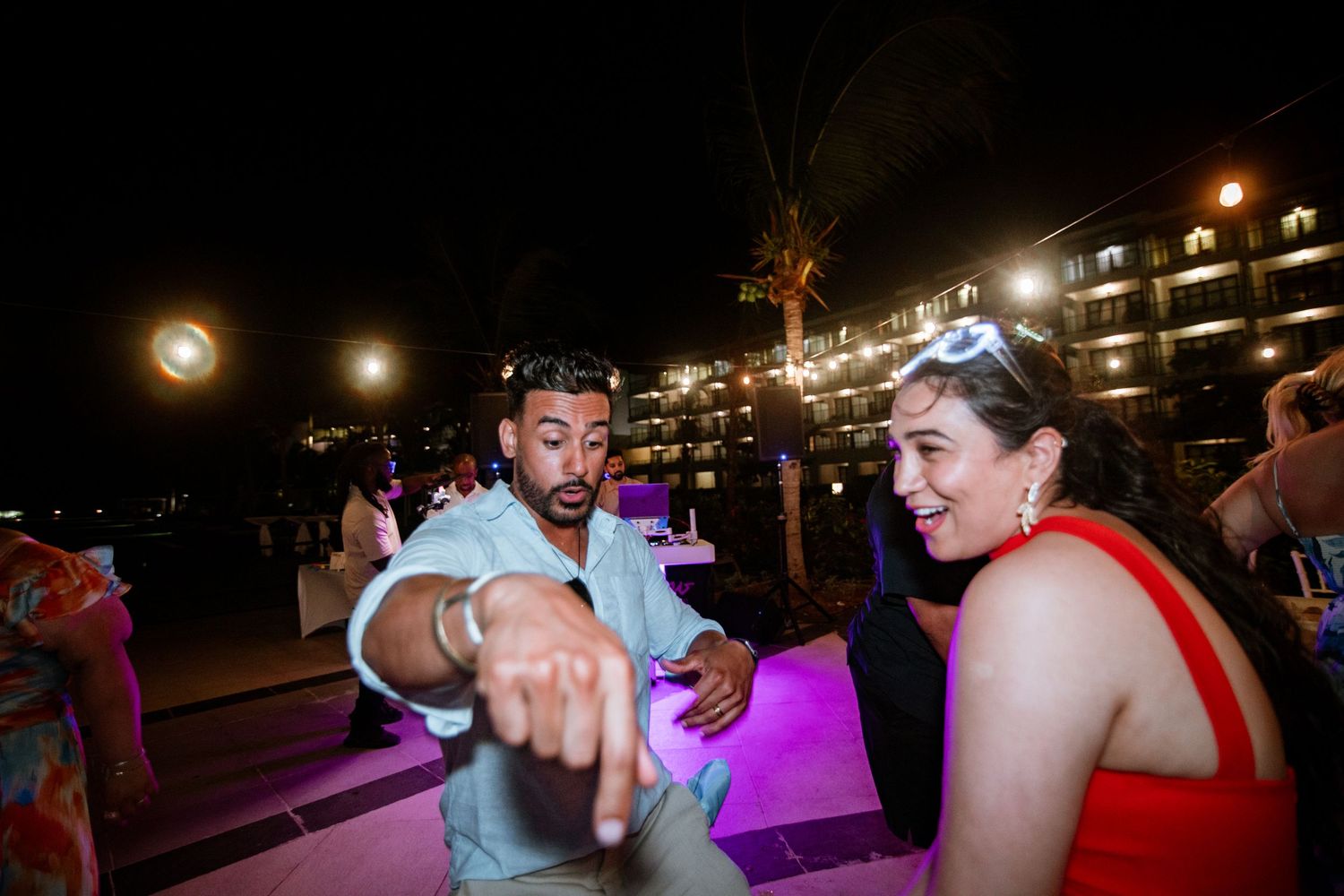 Guests dancing and pointing at colorful outdoor nighttime wedding reception by resort pool.