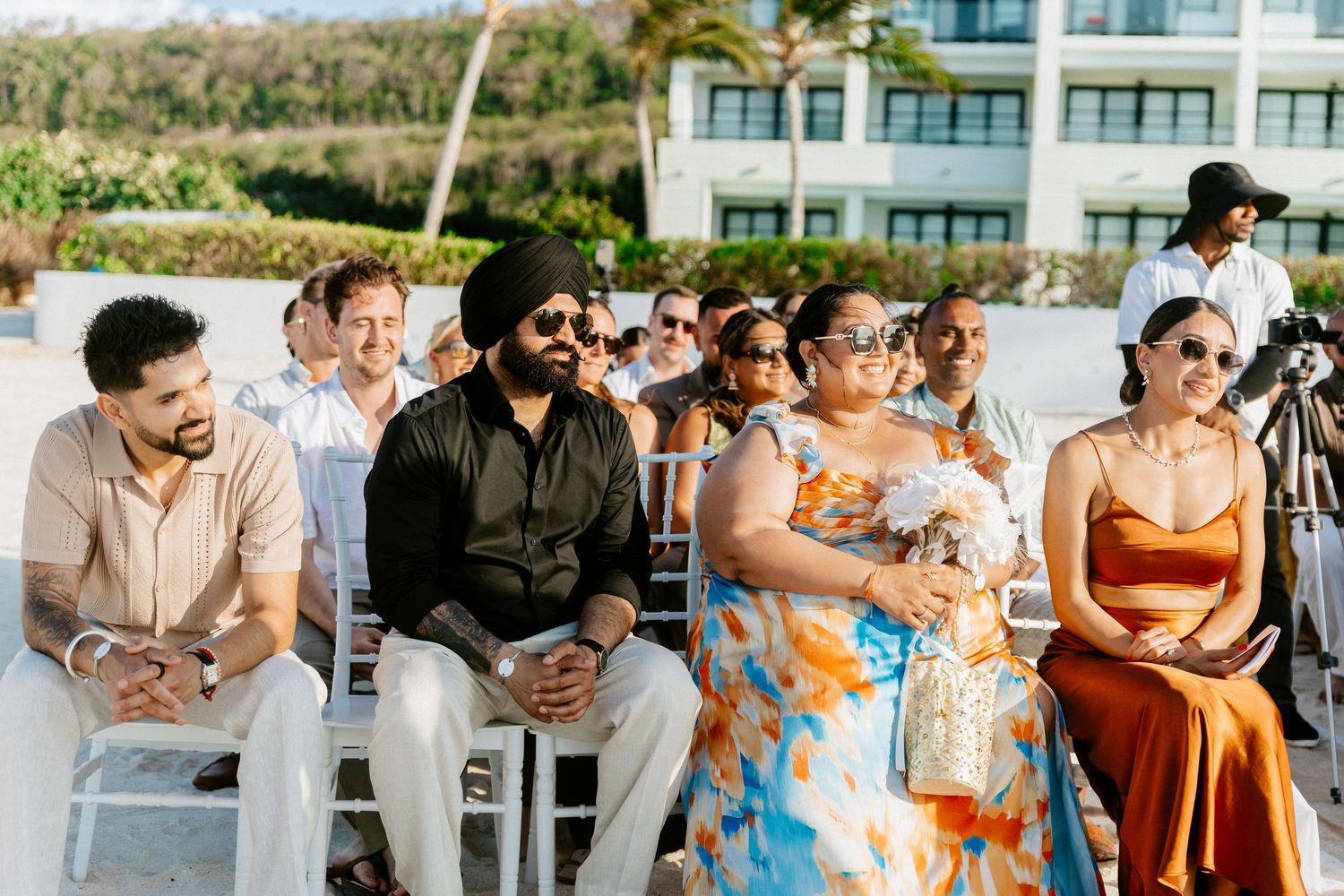 Wedding guests seated outdoors in white chairs watching beach ceremony on sunny day.