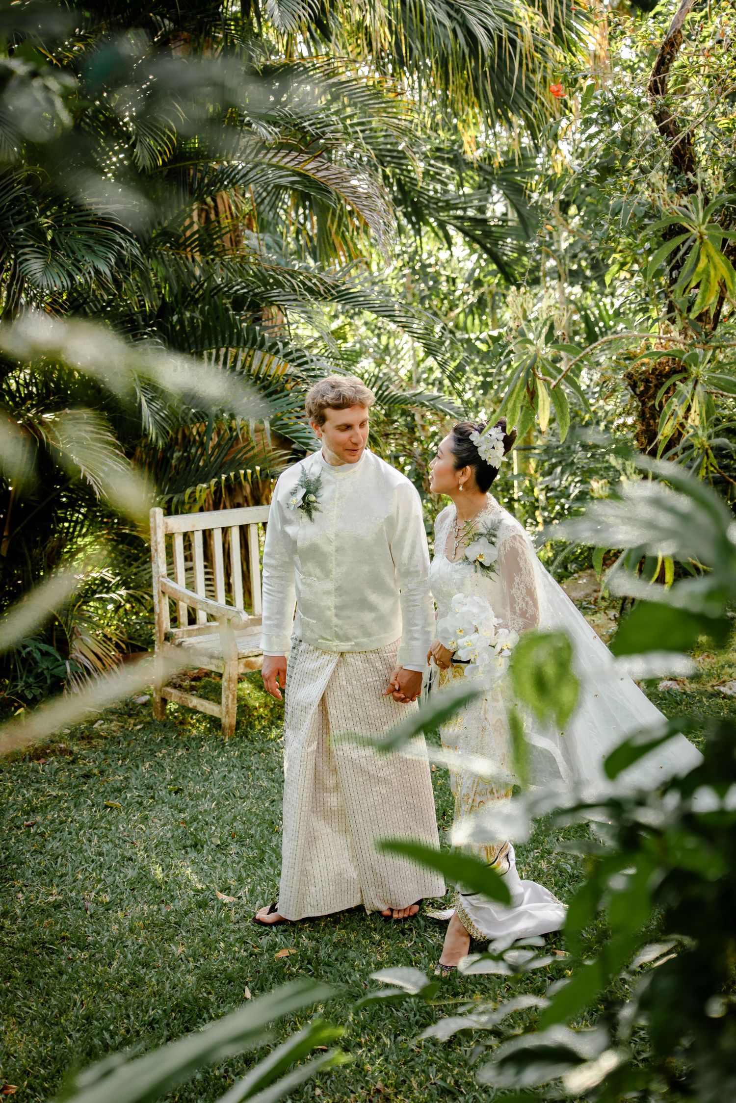 Couple in white traditional wedding attire holding hands in a lush tropical garden with palm trees.