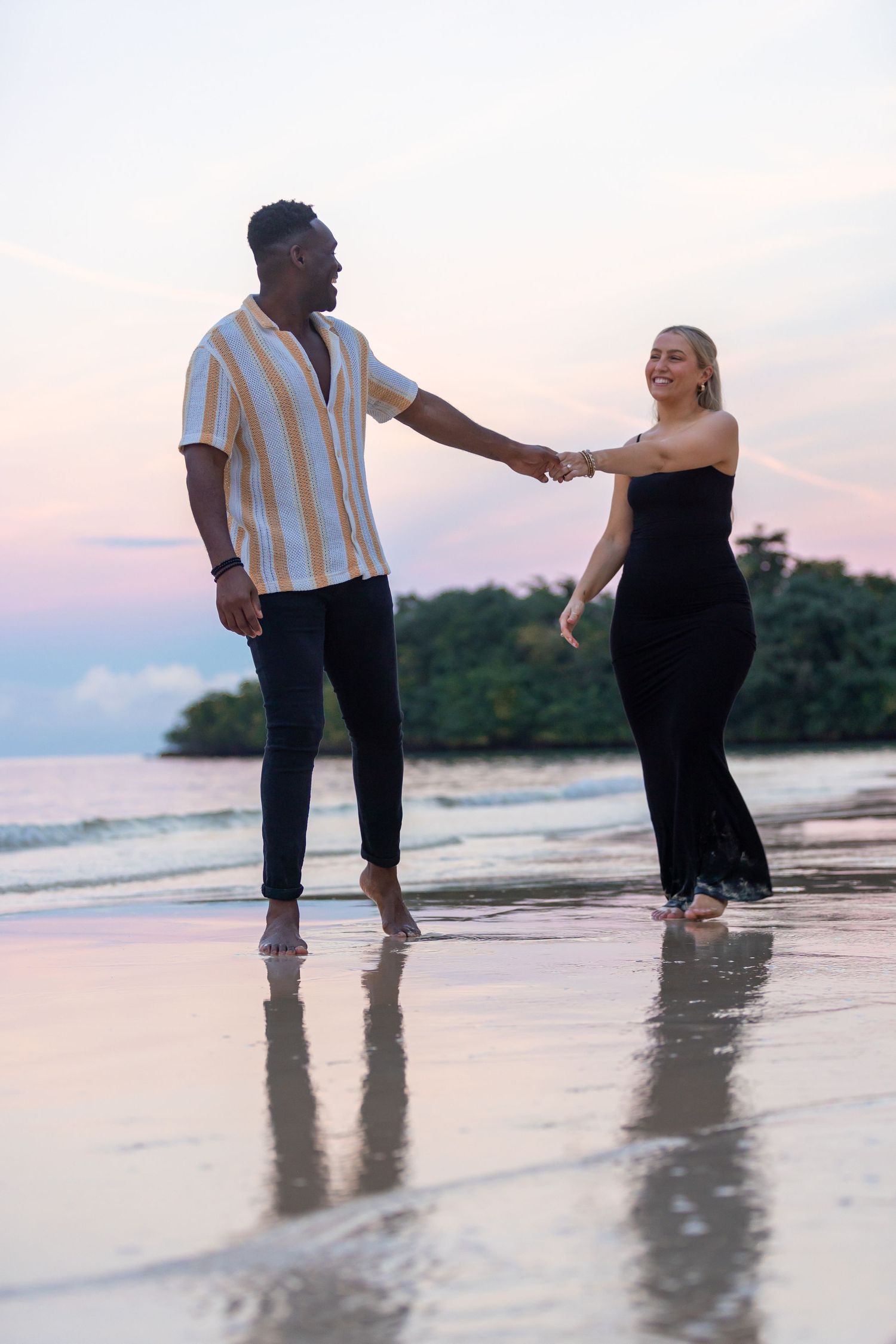 Playful interracial couple holding hands on a reflective beach with pink sunset sky.