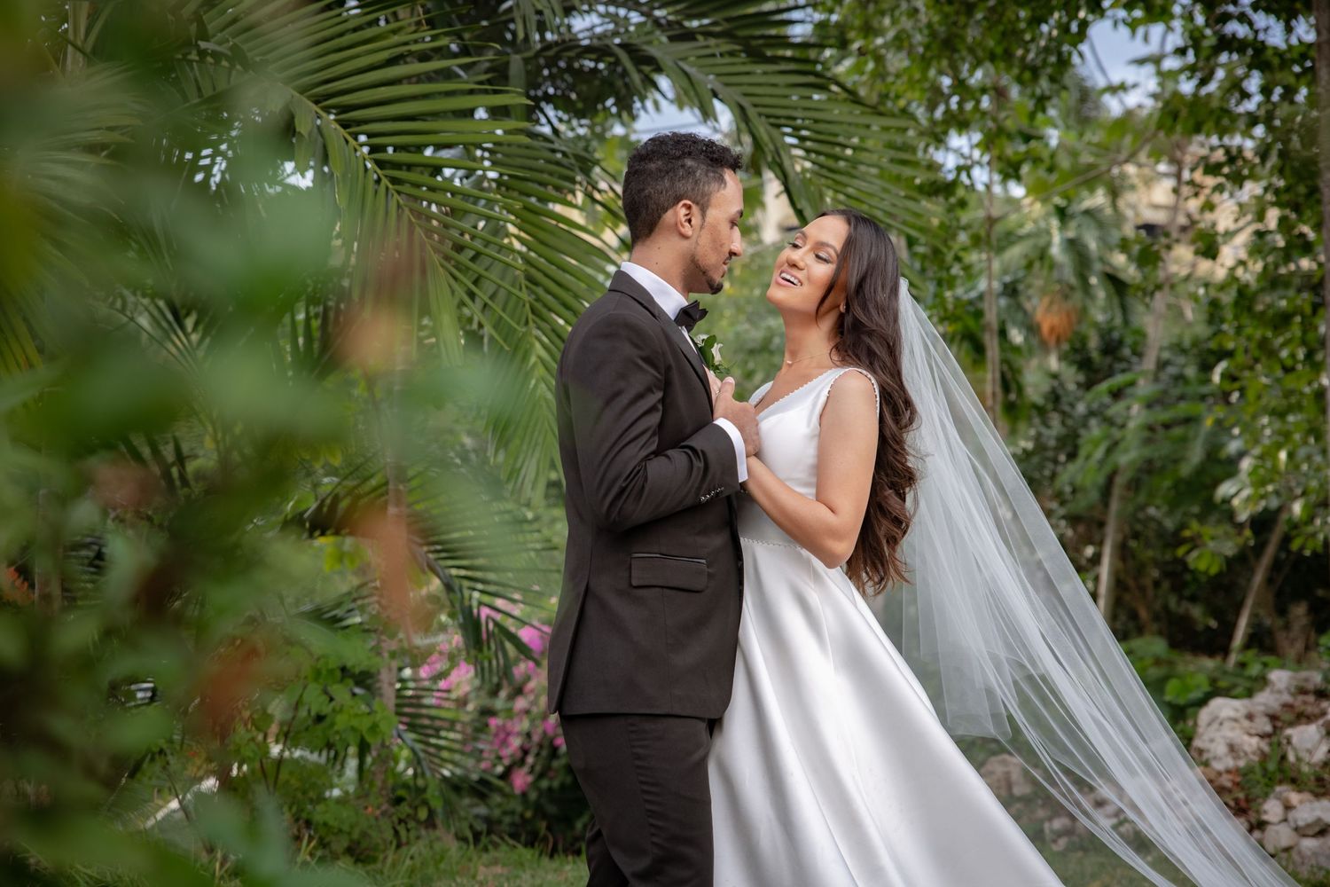 Bride and groom sharing romantic moment surrounded by tropical greenery and pink flowers.