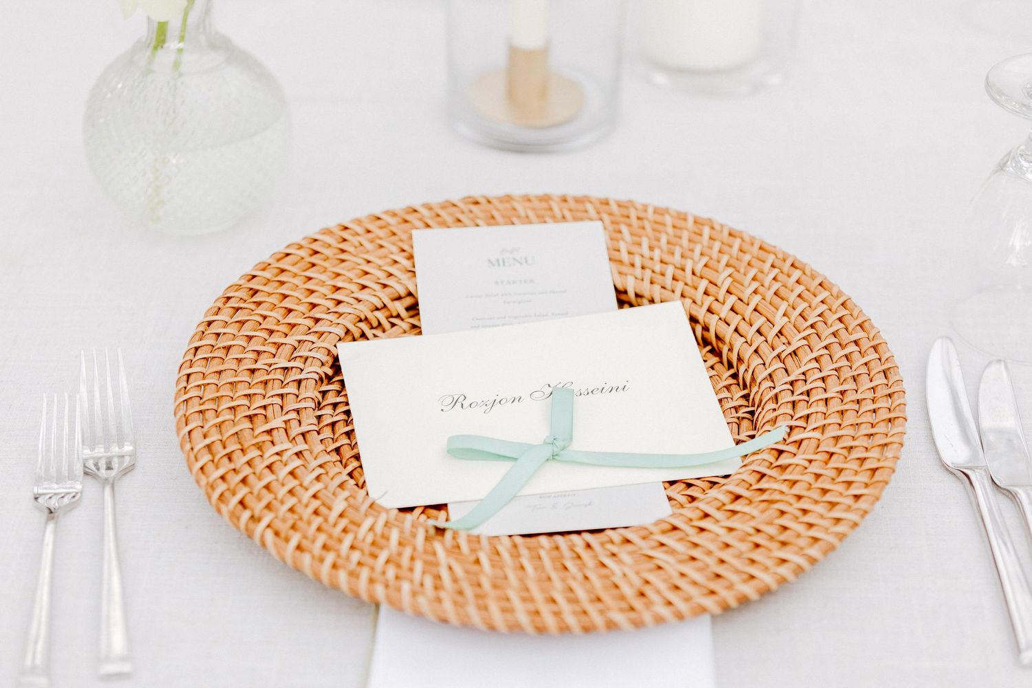 Elegant wedding place setting with woven rattan charger plate, mint ribbon-tied menu card on white linen tablecloth.