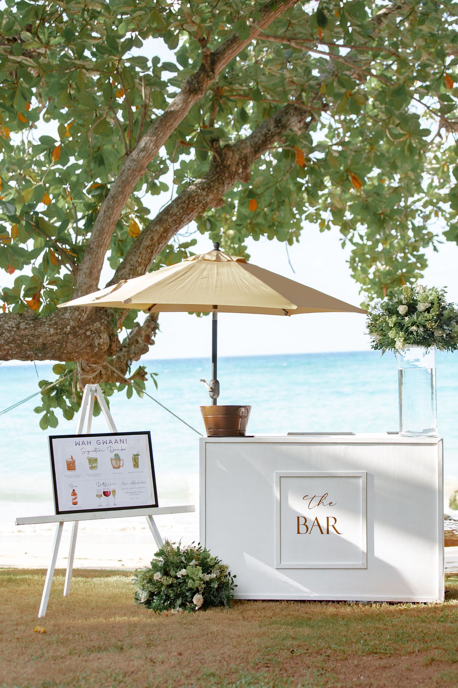Elegant beachside wedding bar setup with white counter, umbrella, and tropical greenery near turquoise ocean waters.