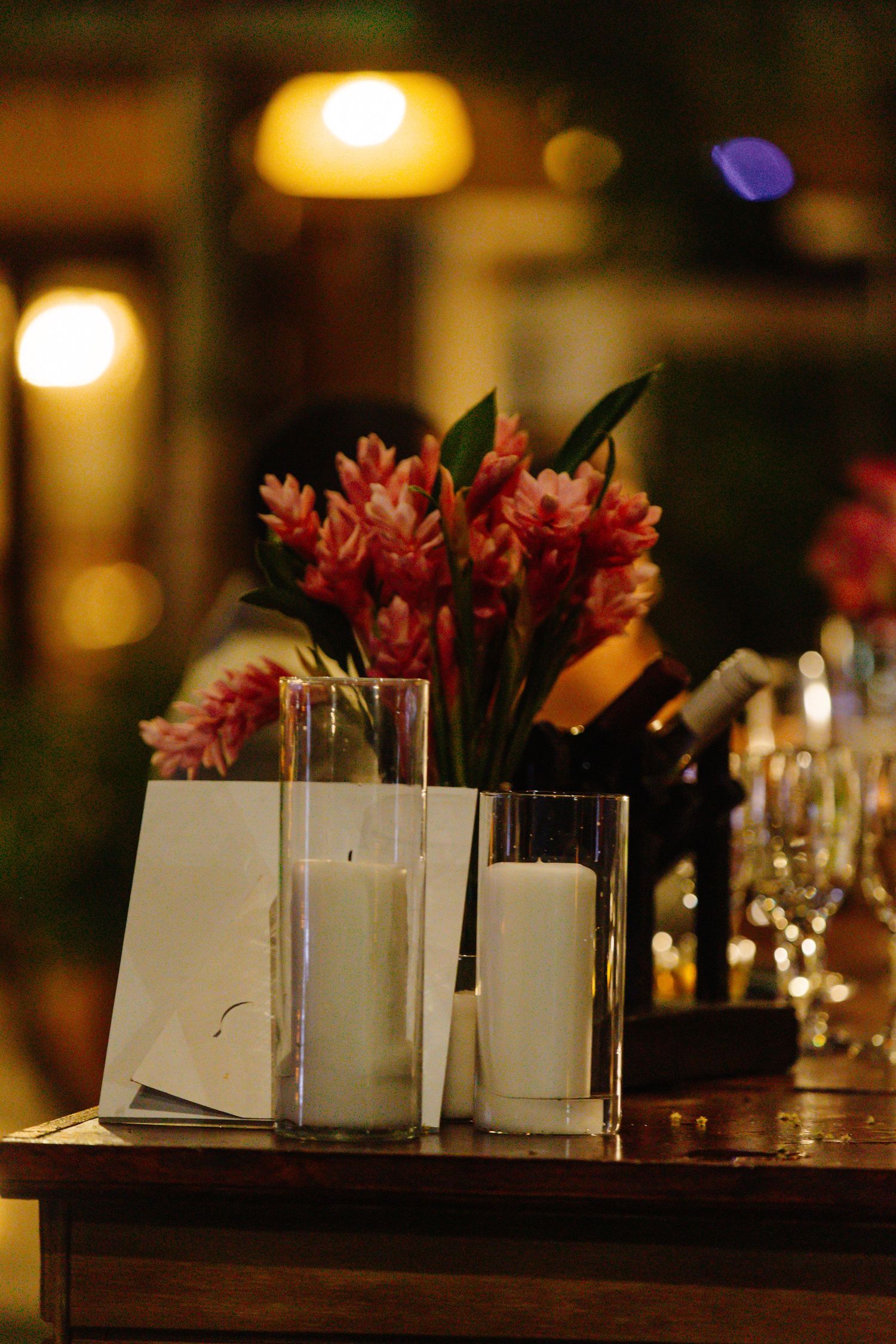 Romantic wedding table decor with white candles in glass holders and pink tropical flowers on a wooden surface.