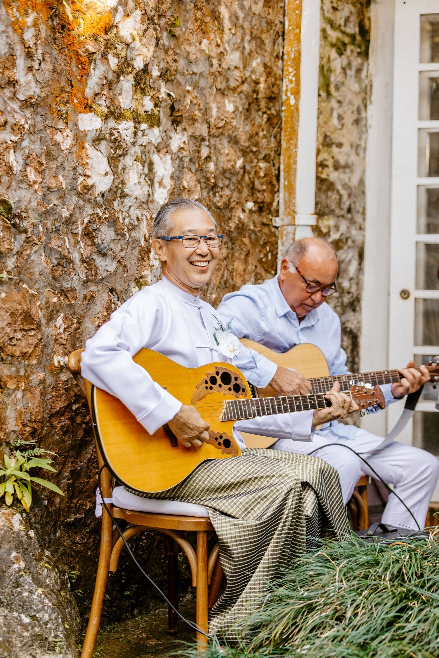 Two men in traditional Myanmar attire playing acoustic guitars and smiling outdoors.