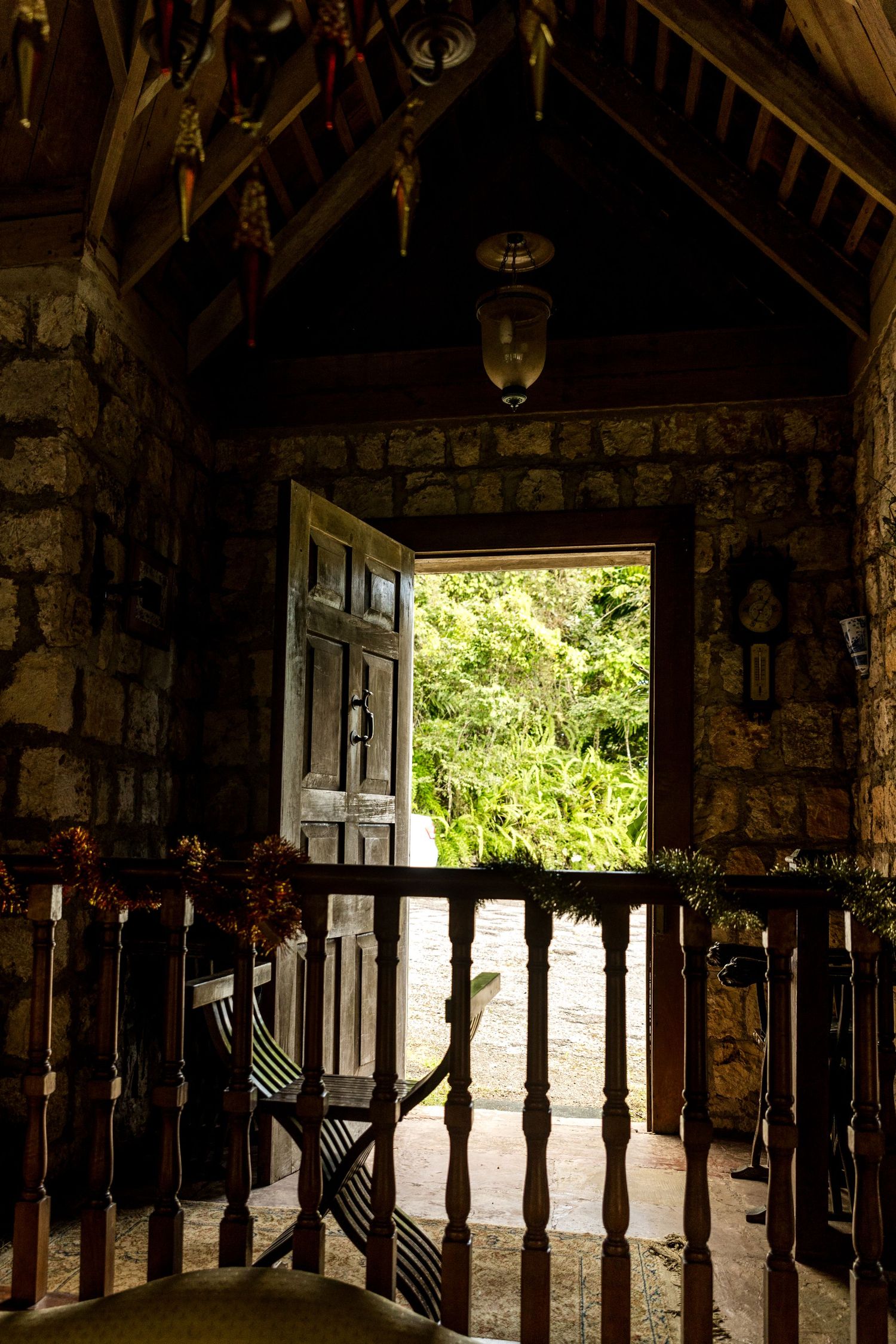 Dark rustic stone chapel interior with wooden door open to lush green garden, featuring ornate ceiling and hanging lantern.