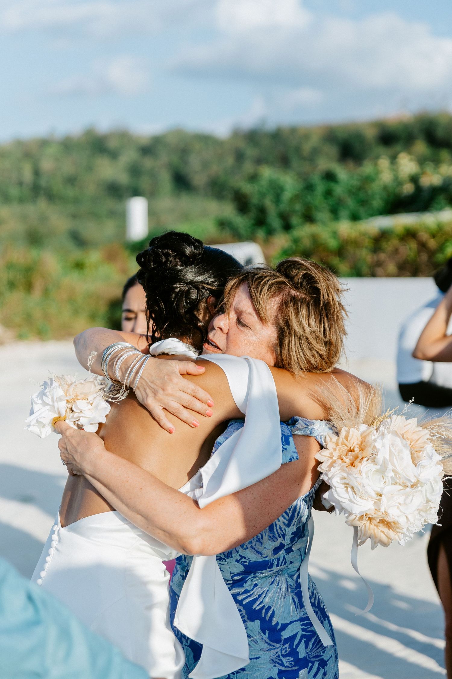 Bride in white backless gown sharing emotional hug with guest at outdoor wedding ceremony.