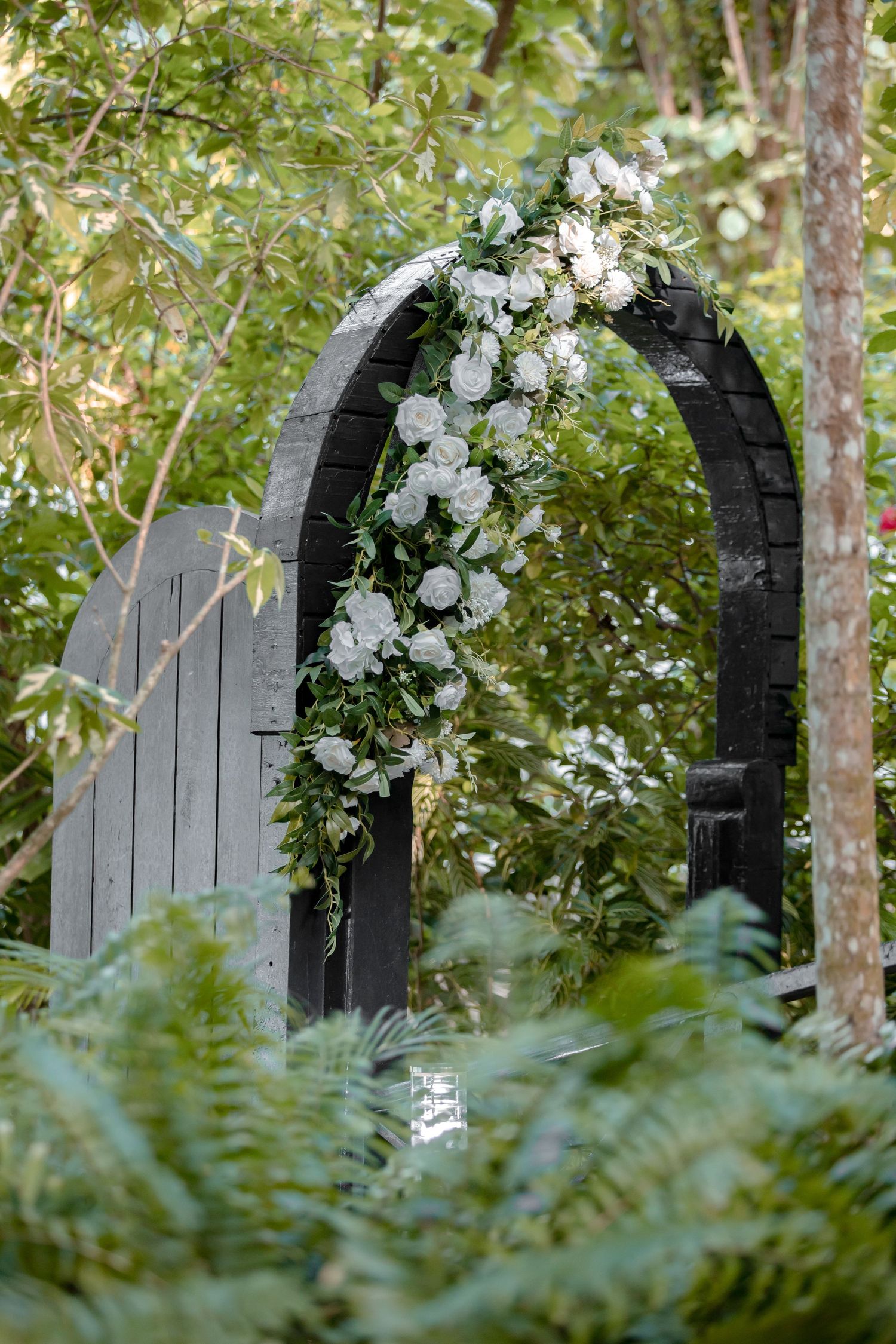 Black wooden arch adorned with white roses and greenery in a lush garden wedding ceremony setting.
