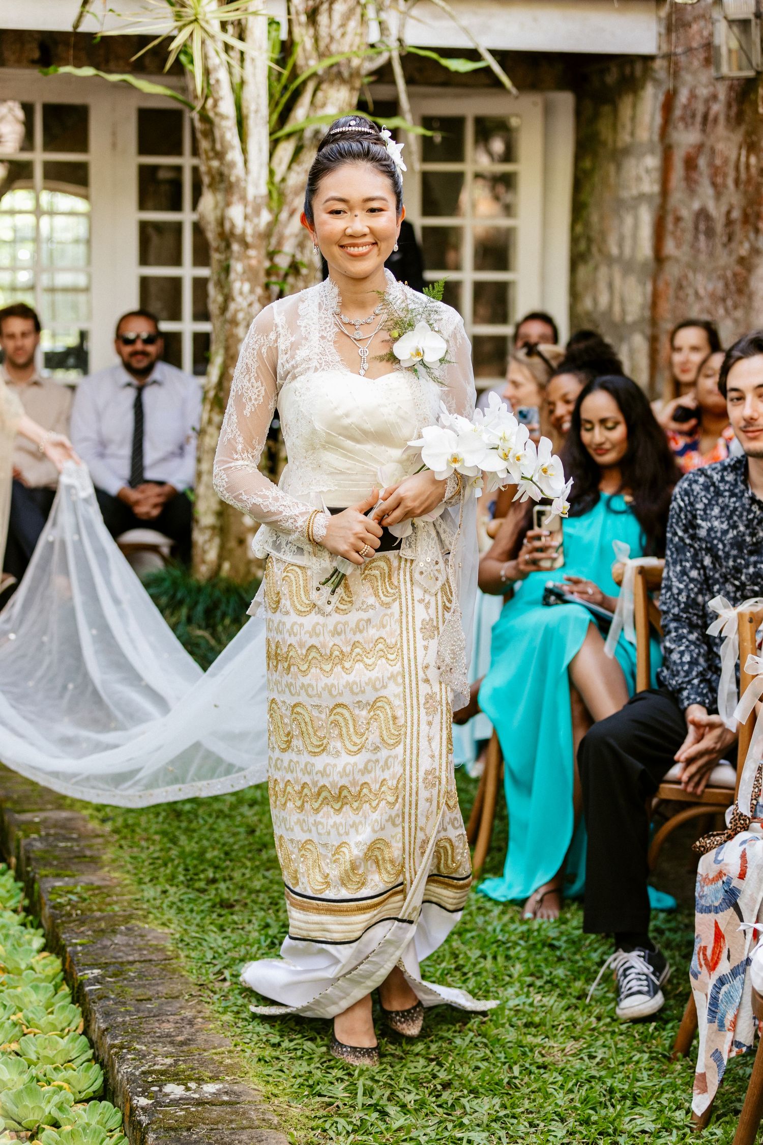 Radiant bride in ornate traditional Asian wedding attire walks down garden aisle with white bouquet.