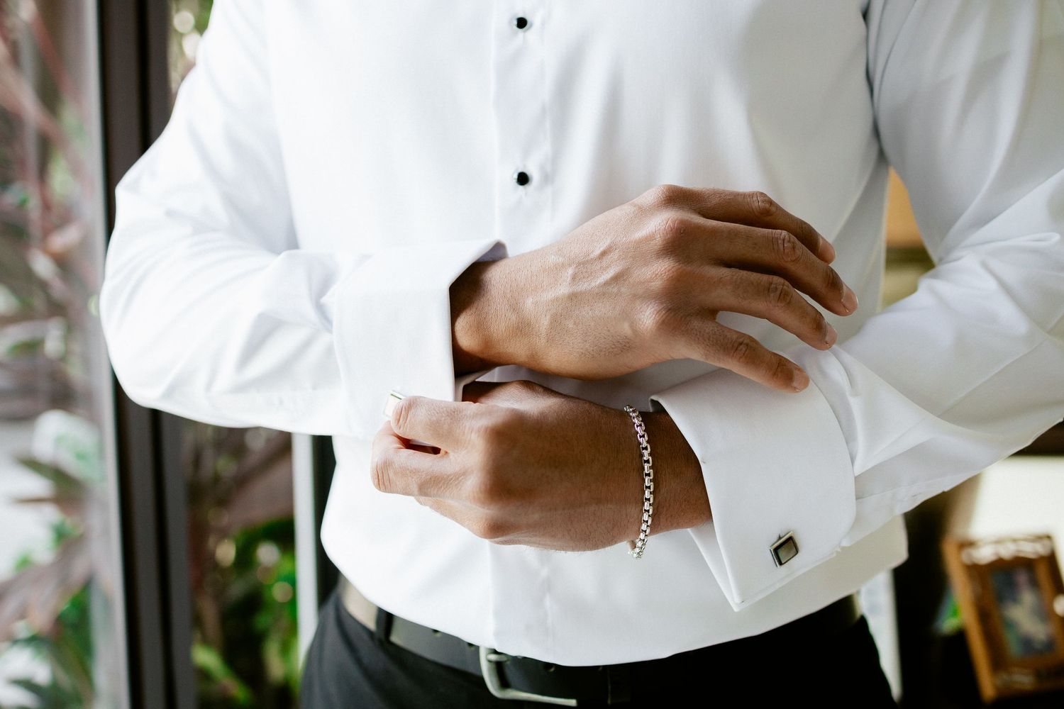A groom in a white tuxedo shirt fastens his cufflinks while wearing a silver bracelet before his wedding.