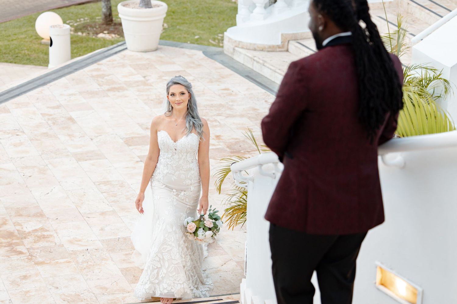 Bride in strapless lace gown walks toward groom during first look at luxury outdoor venue.