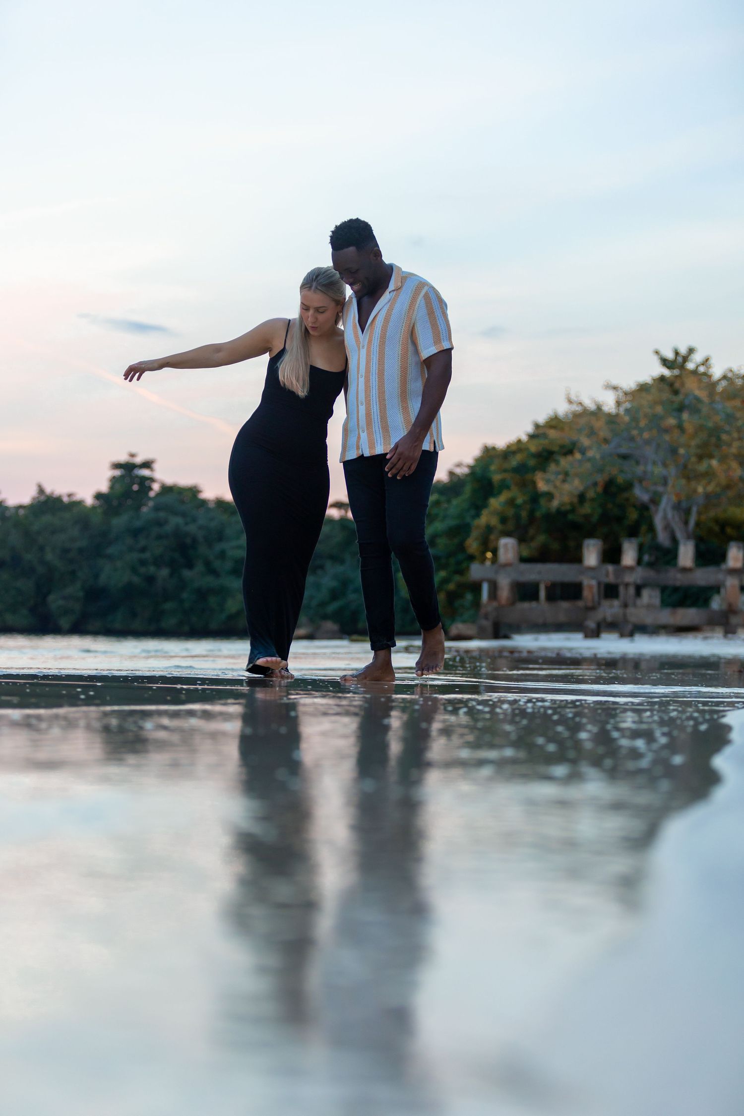 Interracial couple walking barefoot on reflective wet beach at dusk, woman pointing at water.