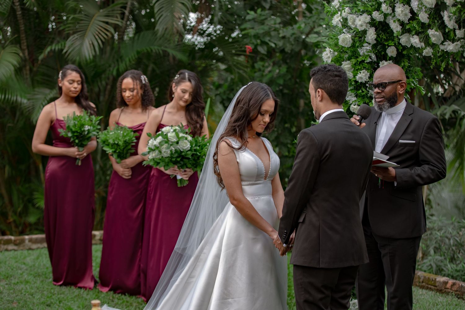 Bride and groom holding hands during outdoor ceremony with bridesmaids in burgundy dresses.