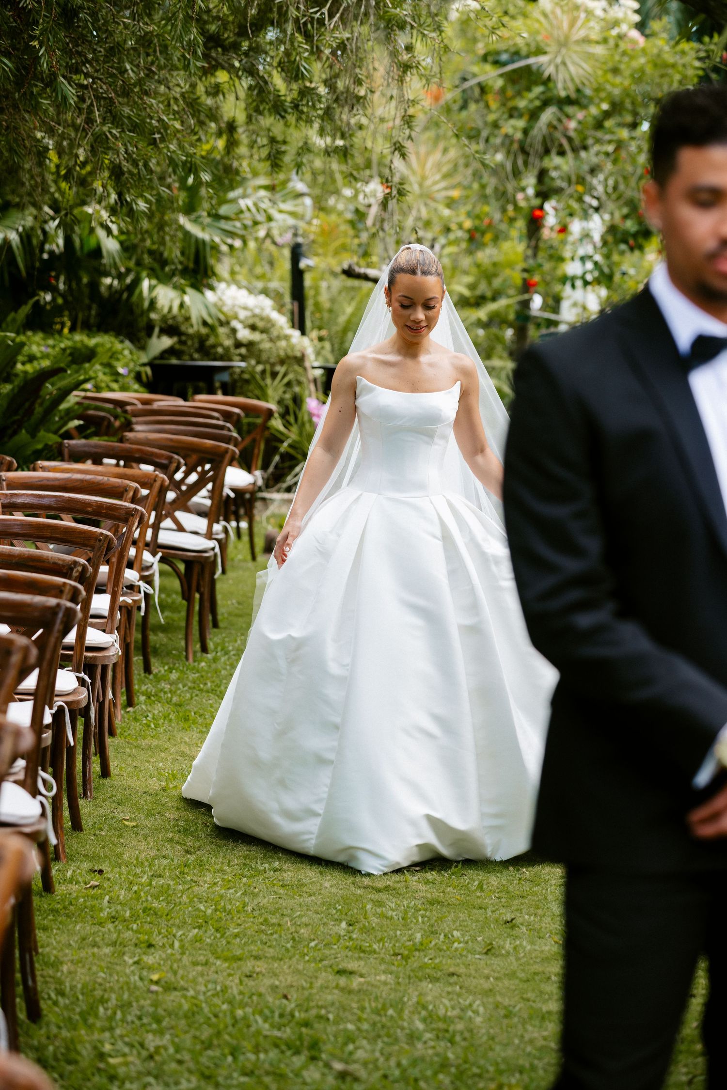 A smiling bride in a strapless white ball gown and veil walks down a lush outdoor wedding ceremony aisle.