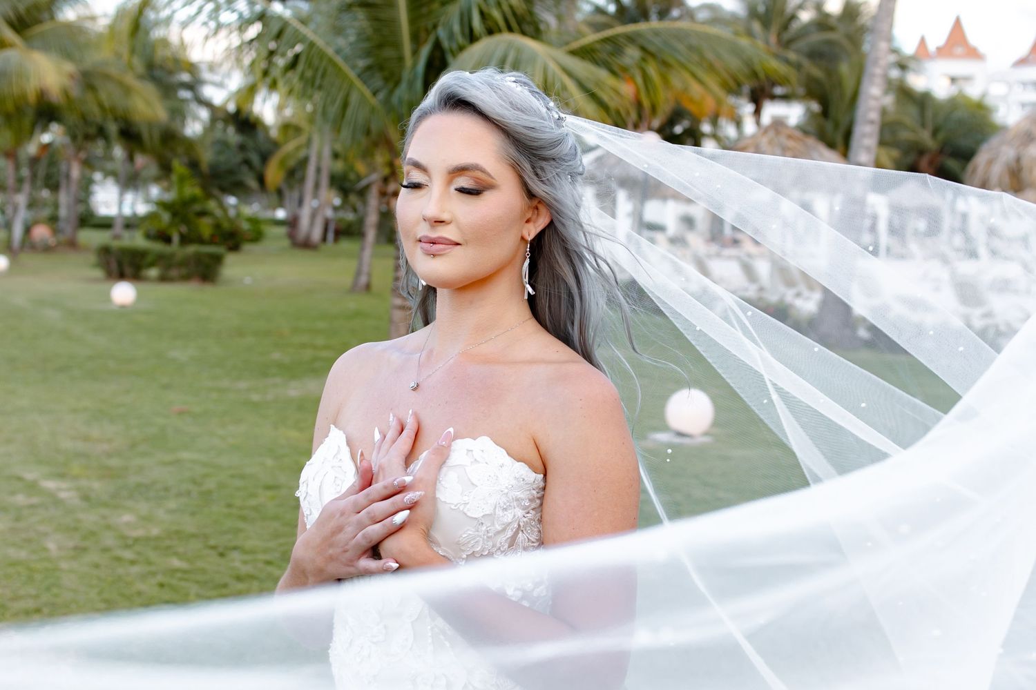 Elegant bride in strapless lace gown with flowing veil poses in lush tropical garden setting.