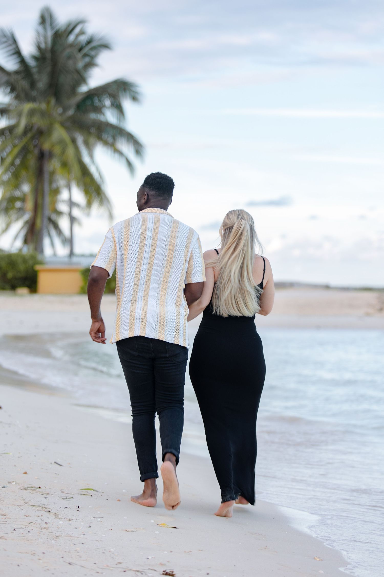 Couple walking barefoot along tropical beach shoreline with palm trees, viewed from behind.