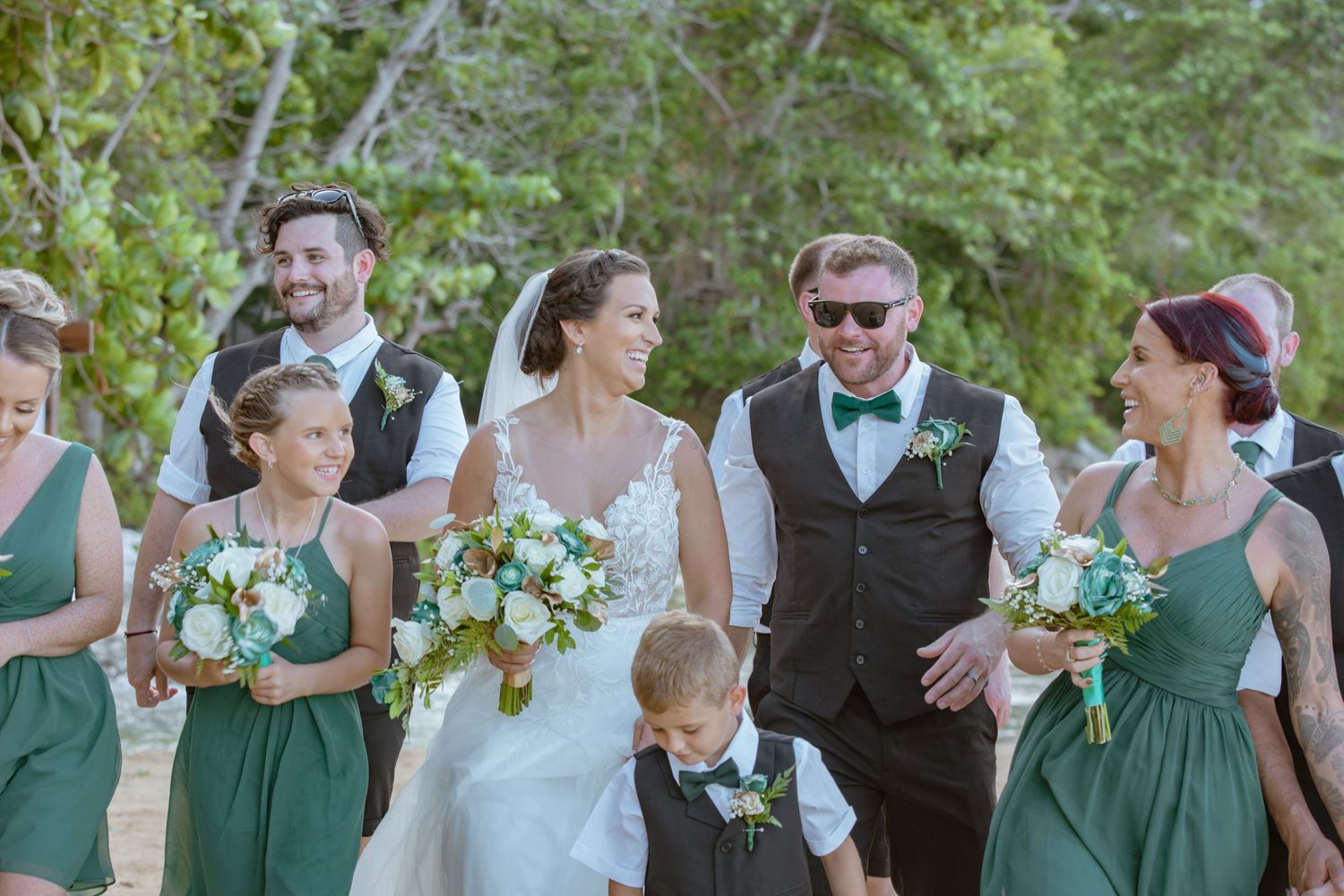 Bride and groom walking with wedding party in green dresses and dark vests on beach.