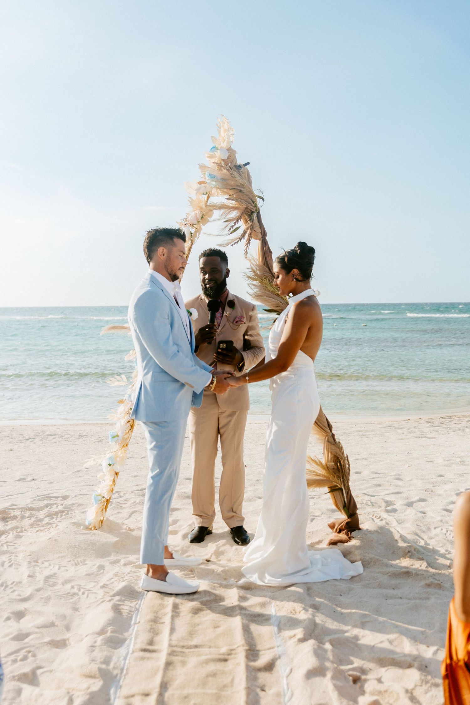 Bride and groom exchange vows on beach with officiant and pampas grass arch backdrop.