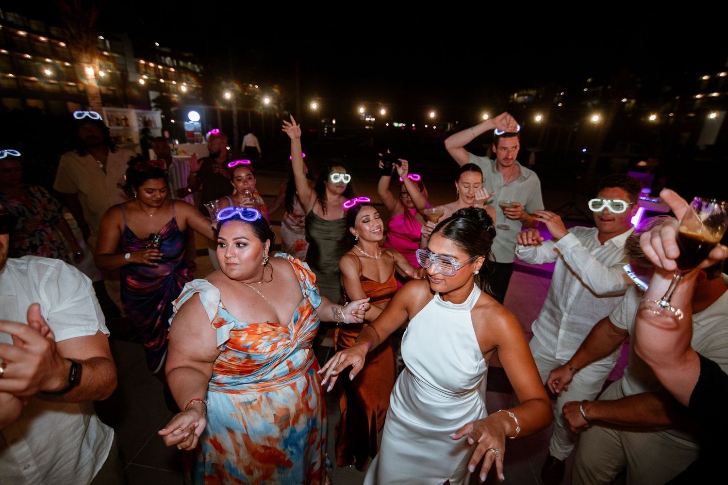 Wedding guests dancing at night outdoor reception wearing neon glow glasses and holding light-up accessories.