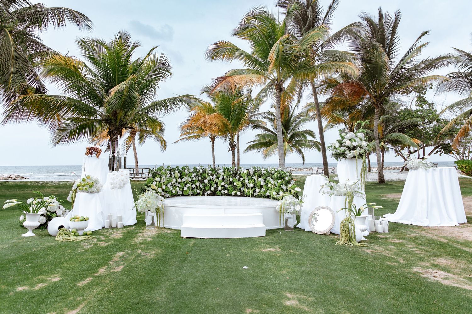 Elegant outdoor wedding ceremony setup with white floral arrangements, draped tables, and palm trees near the ocean.