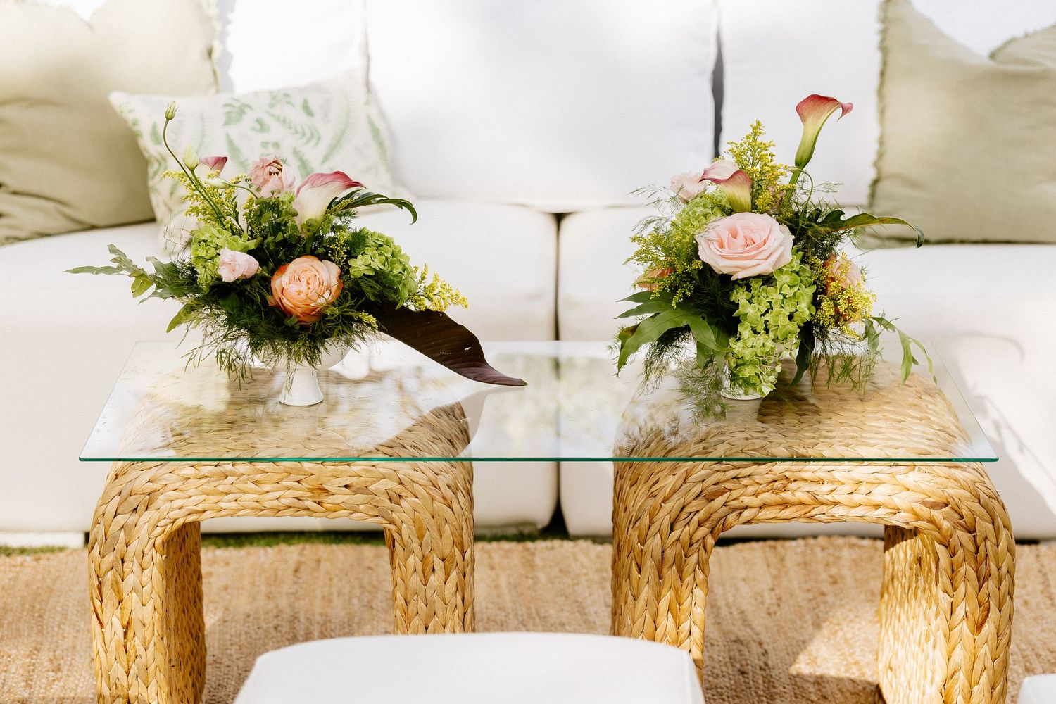 Glass coffee table with floral arrangements on wicker base, beside a white sofa with green cushions.