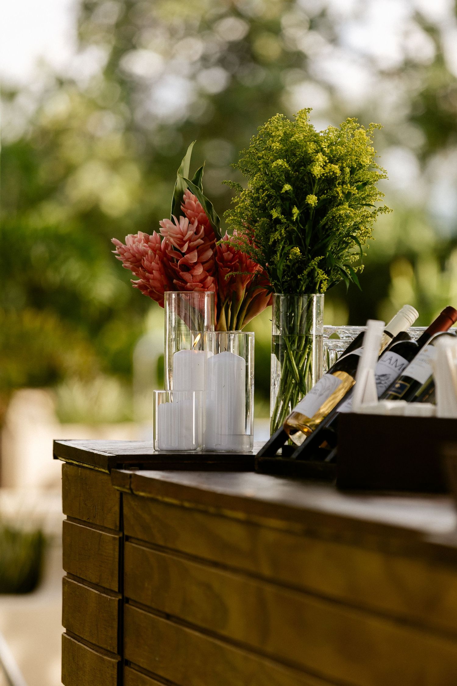 Elegant outdoor bar setup with pink and green floral arrangements in glass vases beside wine bottles on a wooden counter.