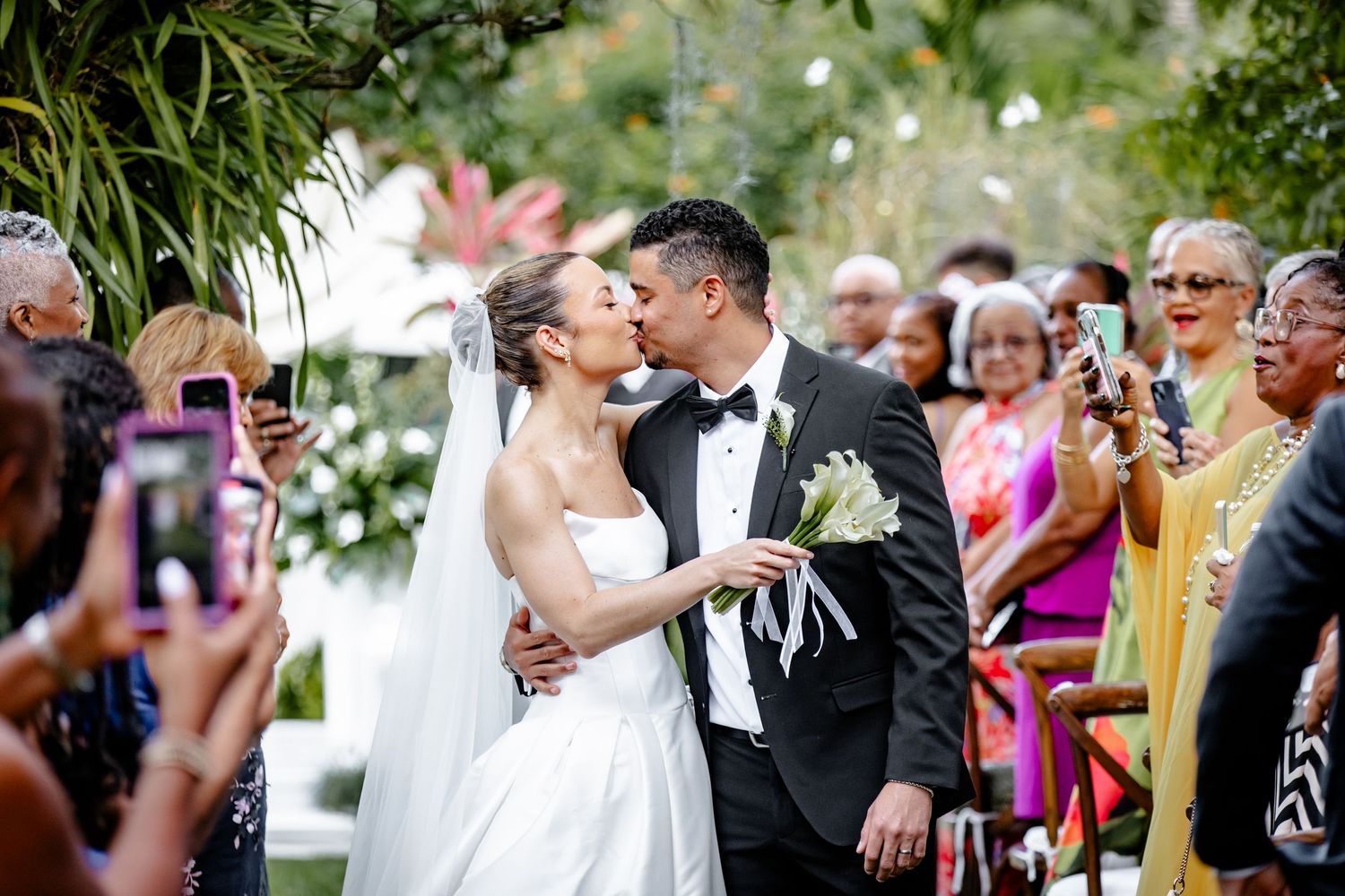 Newlyweds sharing their first kiss as a married couple while guests photograph the joyful moment.