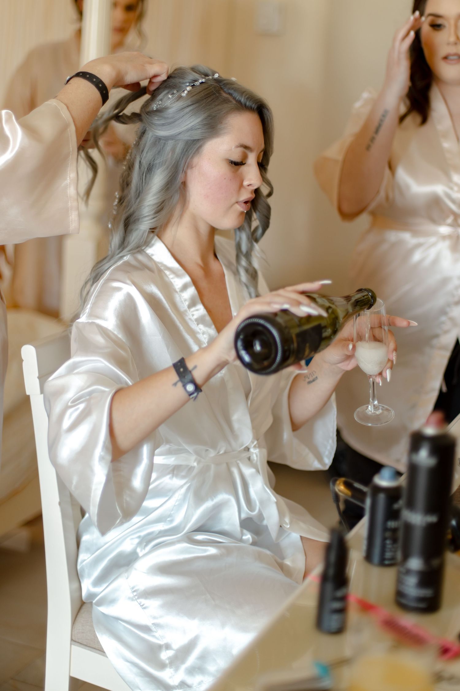 Bride in white satin robe pouring champagne while getting hair done before wedding ceremony.
