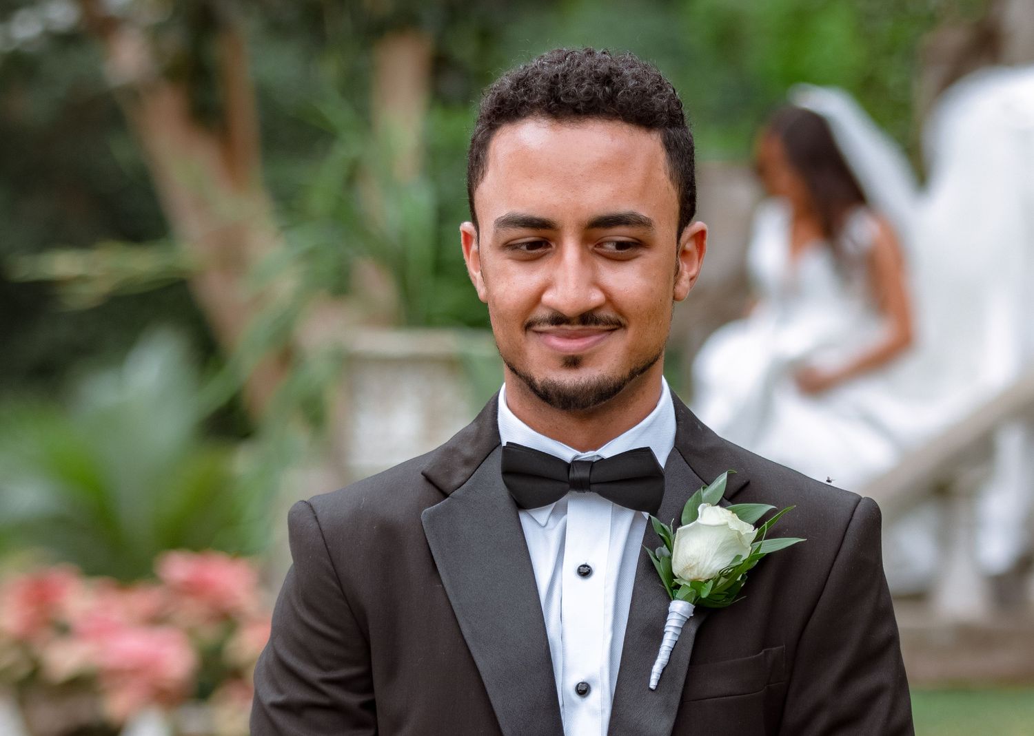 Close-up portrait of smiling groom in black tuxedo with white boutonniere at outdoor wedding.