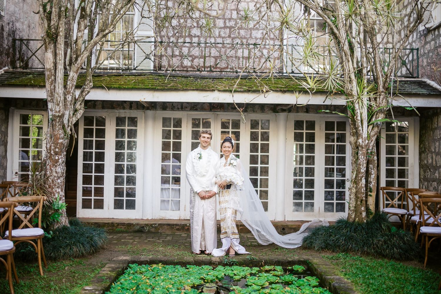 Bride and groom in white wedding attire posing in a lush garden courtyard of a vintage building.