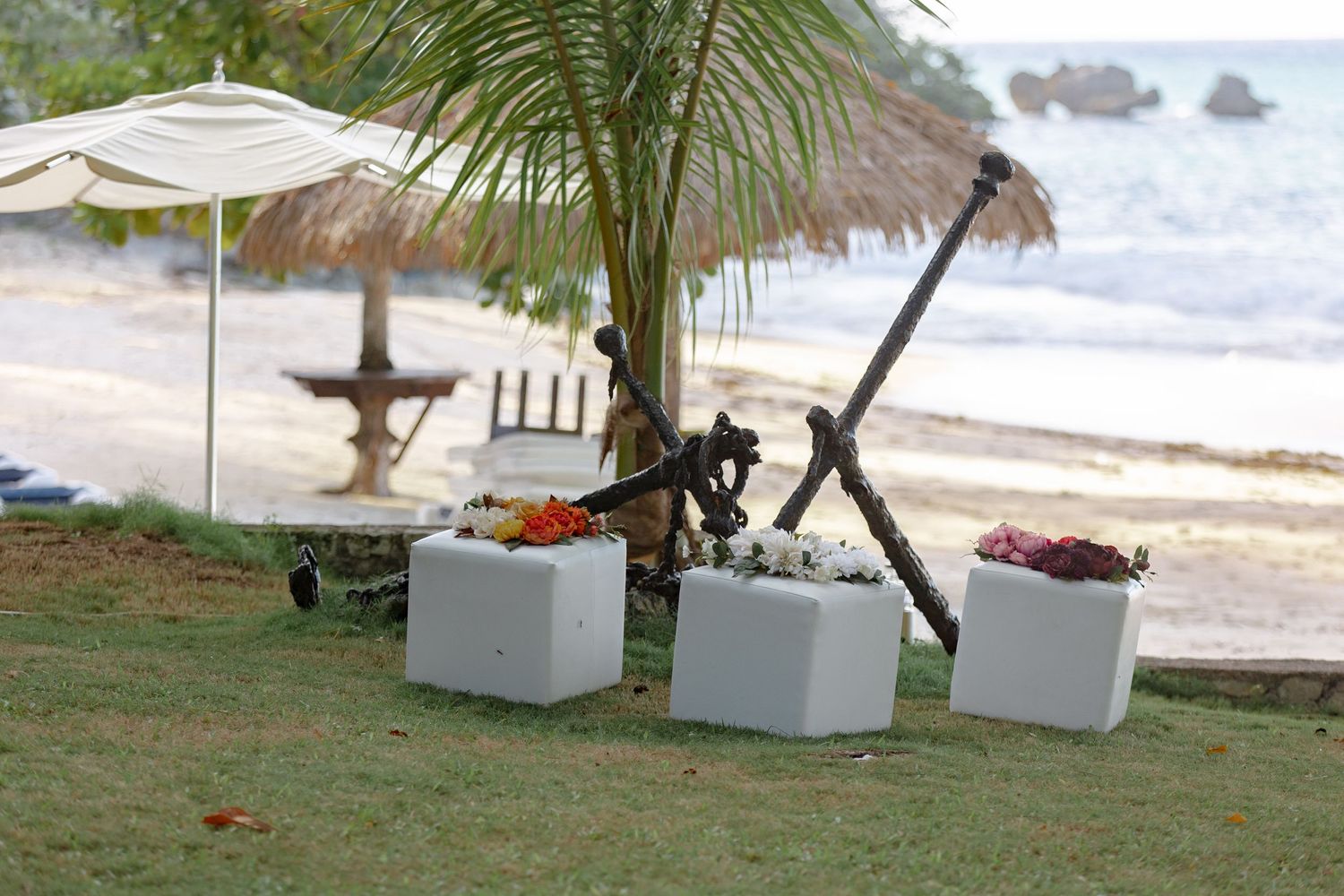 Tropical beach resort with white cube planters, palm tree, anchor decor, and thatched umbrella by the ocean.