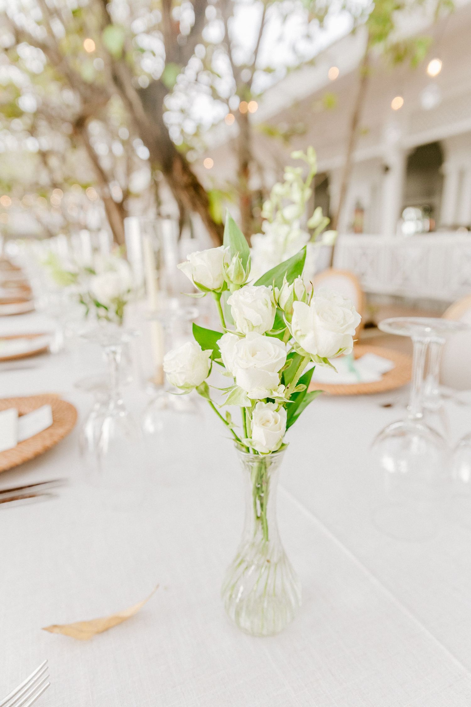 Elegant wedding reception table with a glass bud vase of white roses and green foliage as a minimalist centerpiece.