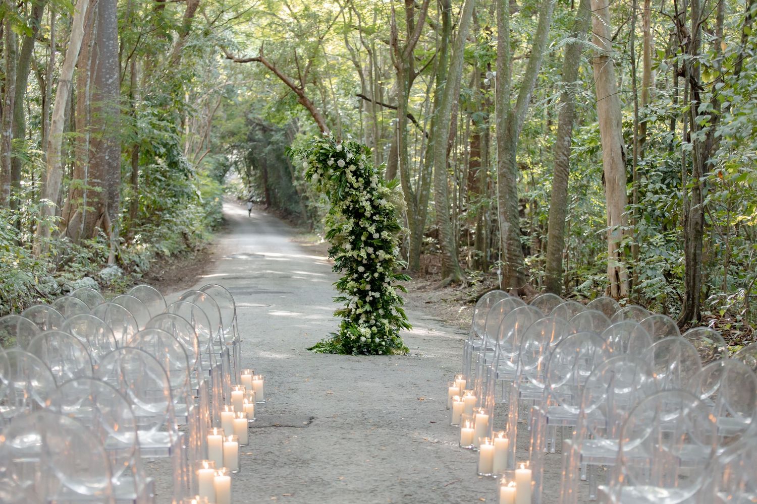 Elegant outdoor wedding ceremony in a lush forest with clear chairs, candles, and a floral arch centerpiece.