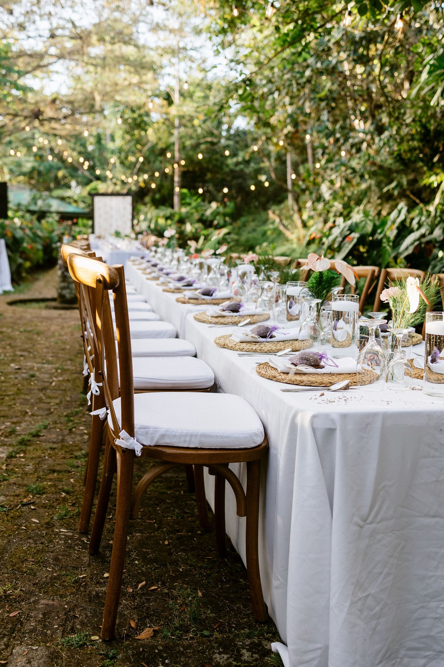 Long outdoor wedding reception table with white linen, woven placemats, and string lights in trees.