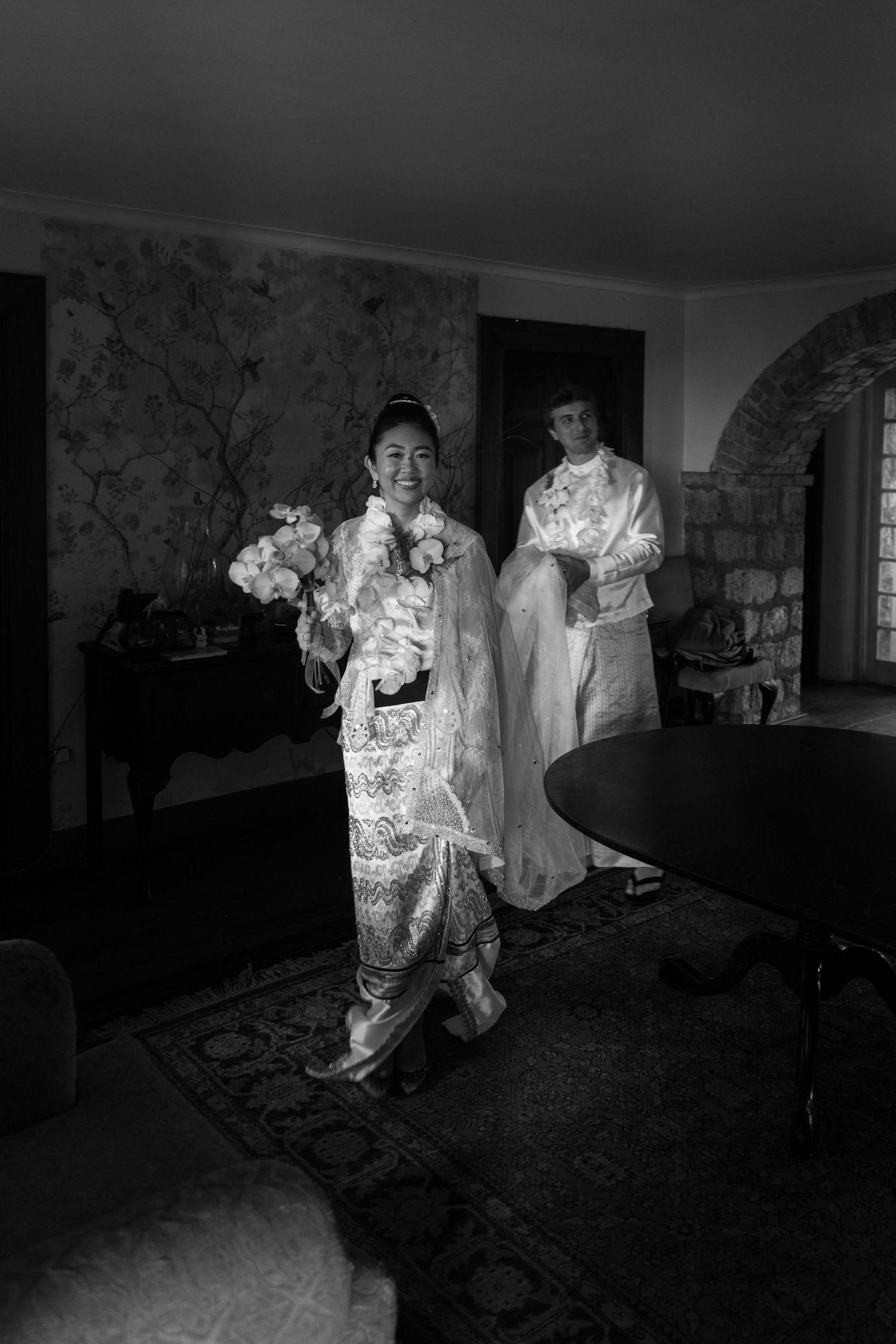 Black and white photo of two women in traditional embroidered gowns holding floral bouquets indoors.
