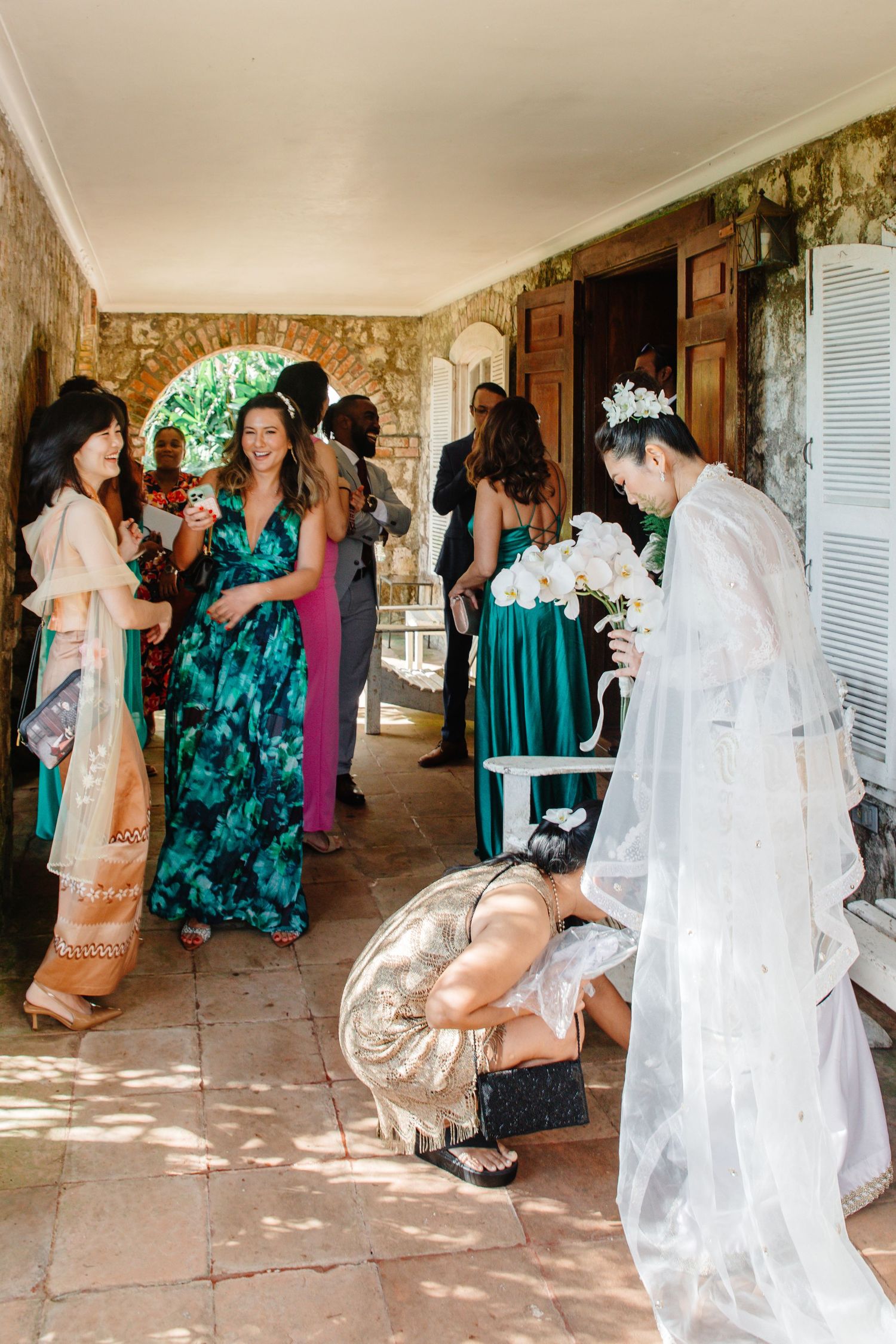 Bride in white gown and veil holding orchid bouquet greeted by guests in colorful dresses at stone venue.