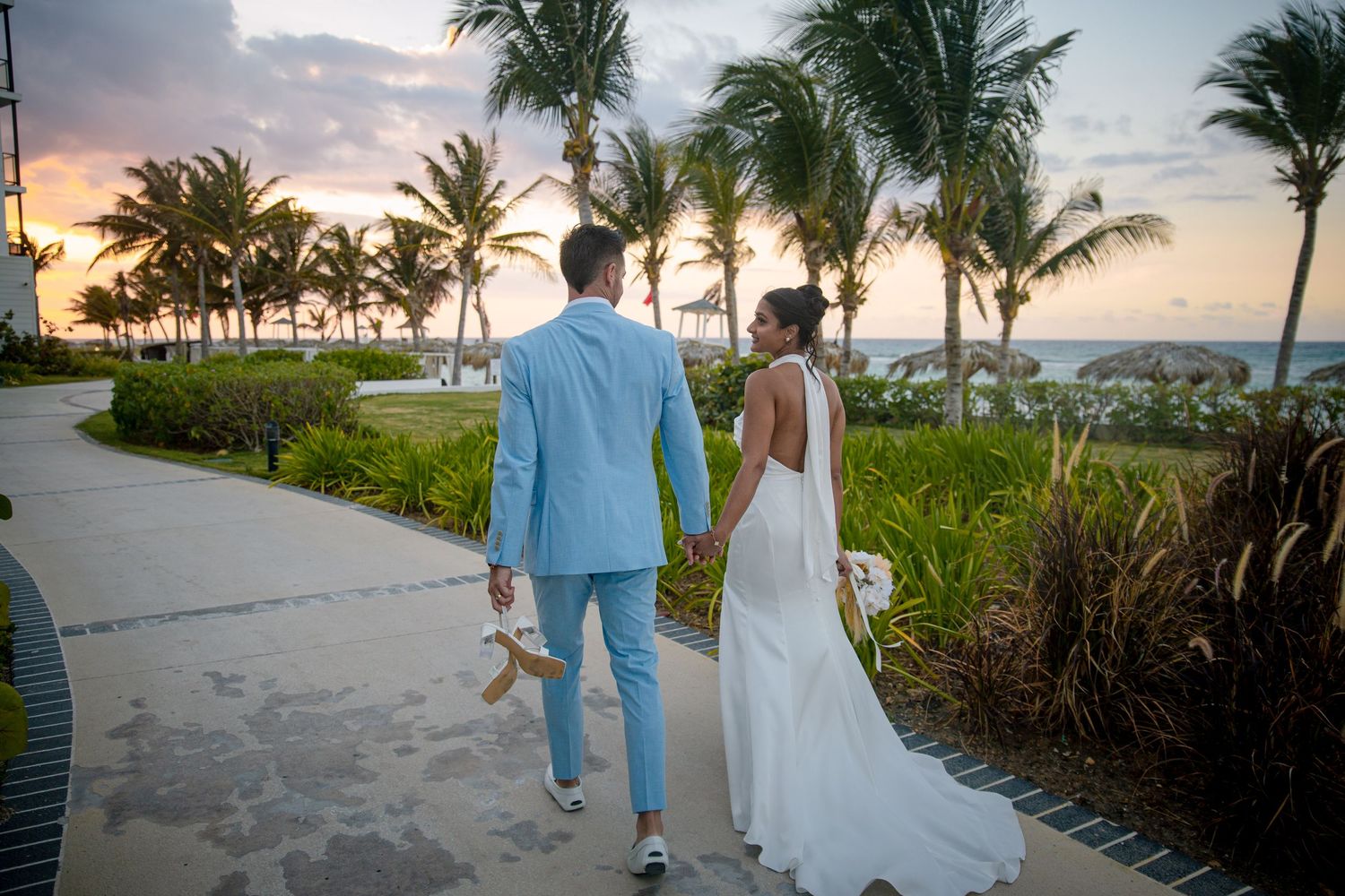 Bride and groom walking hand in hand along tropical resort pathway at sunset.