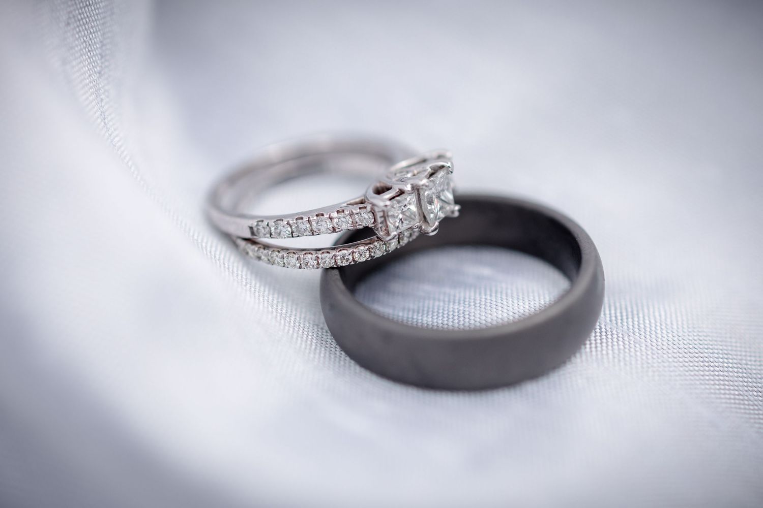 Diamond engagement ring and black wedding band resting on white fabric.