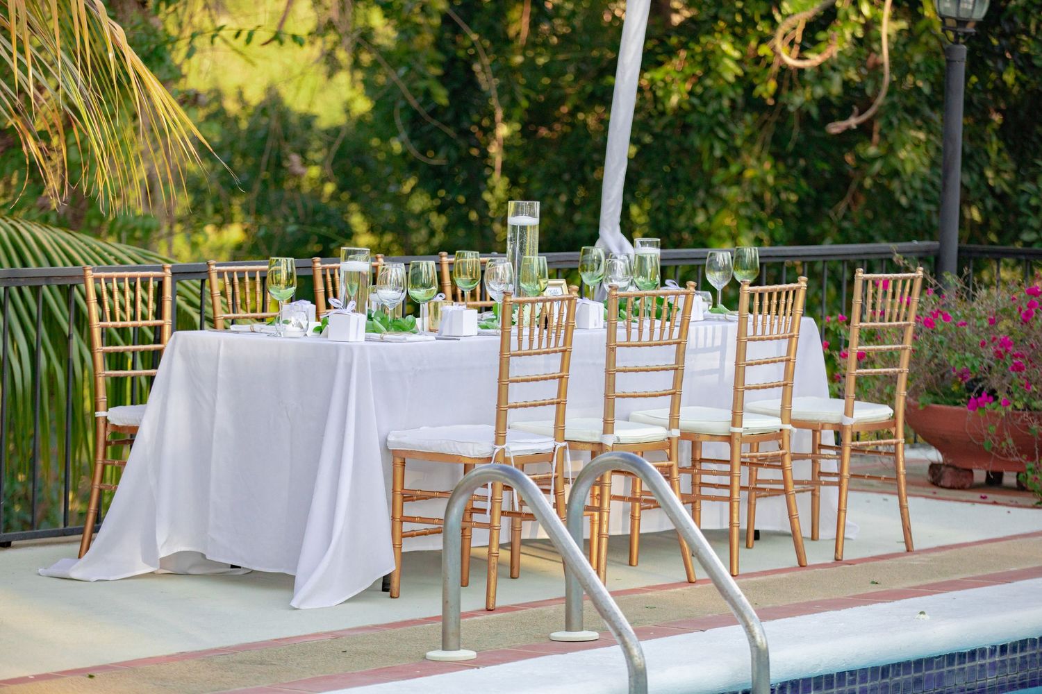Gold chiavari chairs and white linen tables set poolside for an outdoor wedding reception surrounded by tropical greenery.