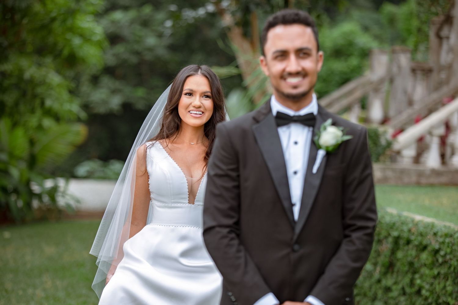 Bride in white gown and veil smiles beside groom in black tuxedo outdoors at wedding.