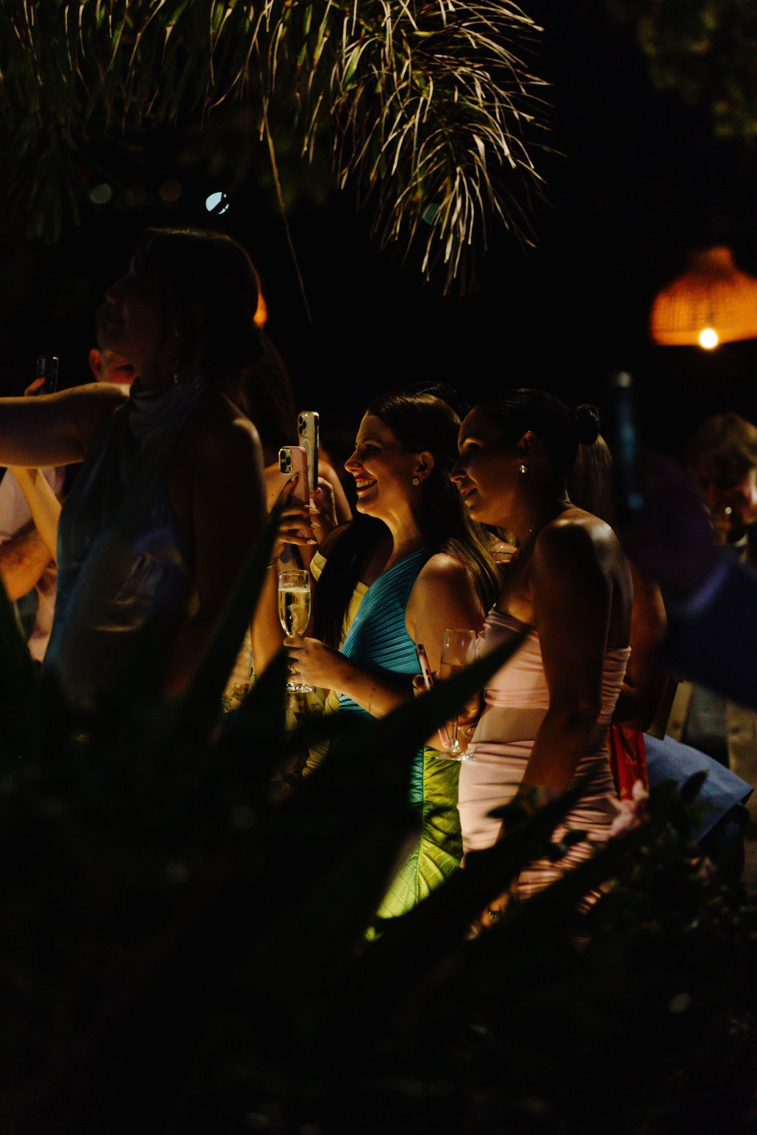 Women in elegant dresses laughing and toasting with champagne at an outdoor evening party.