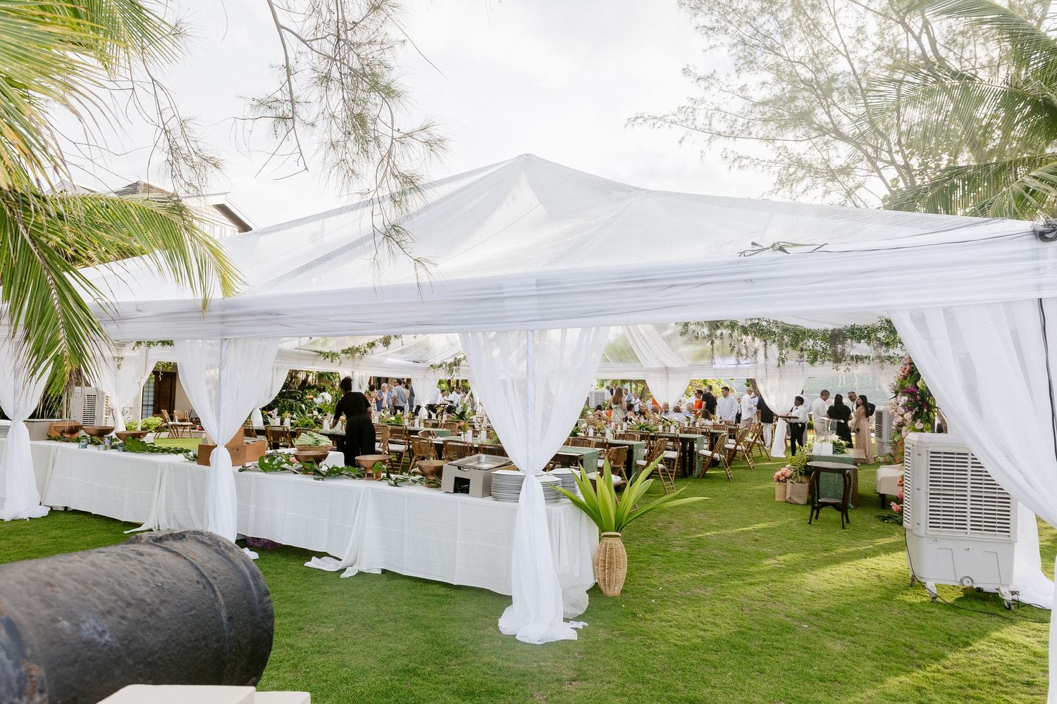 Elegant white outdoor wedding tent with draping curtains, lush greenery, and guests dining on a tropical lawn.