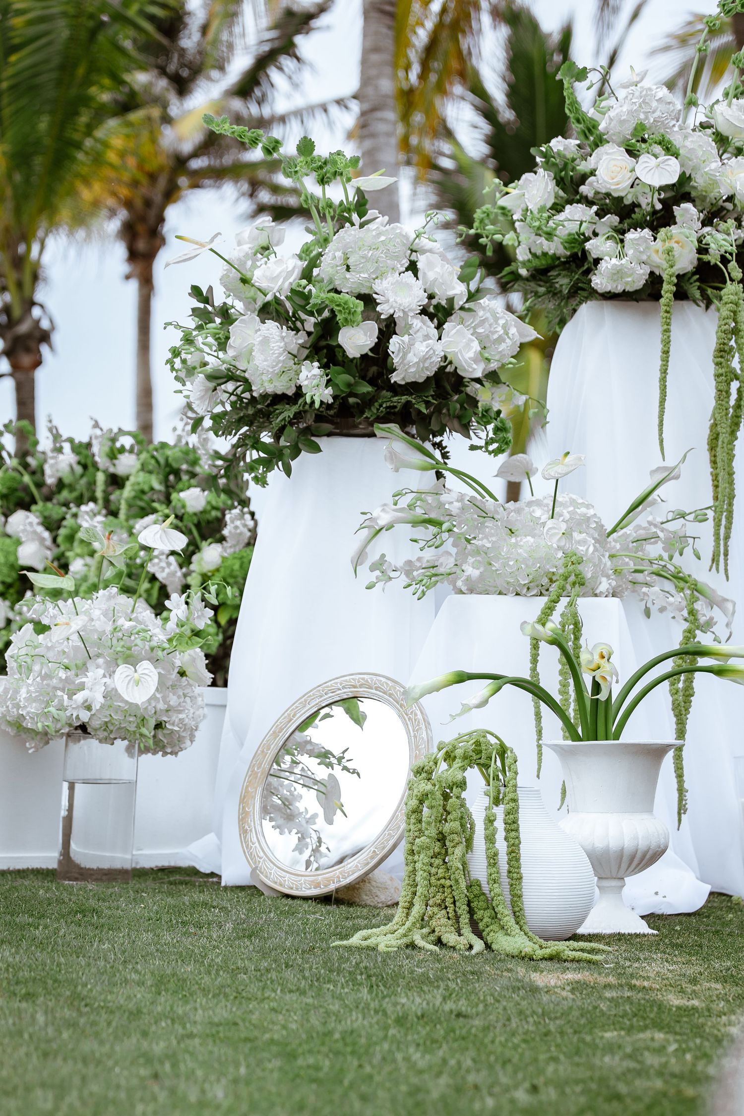 Elegant outdoor wedding decor with white floral arrangements, mirror, and tropical greenery.