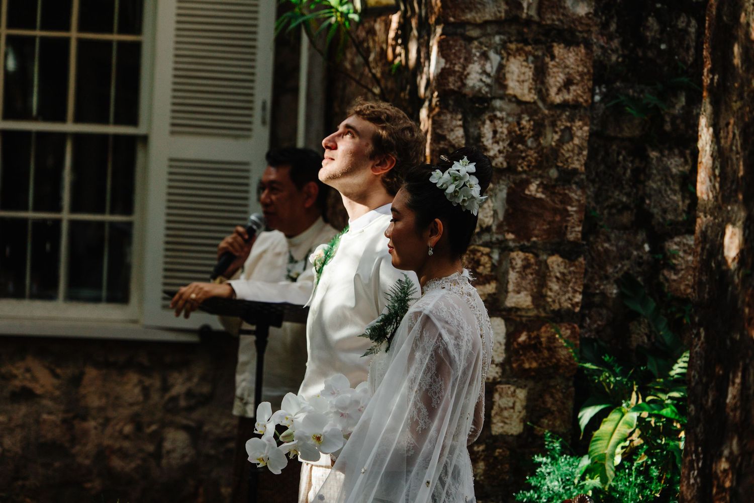 A bride in a white gown and floral veil stands outdoors near a tree during a wedding ceremony.