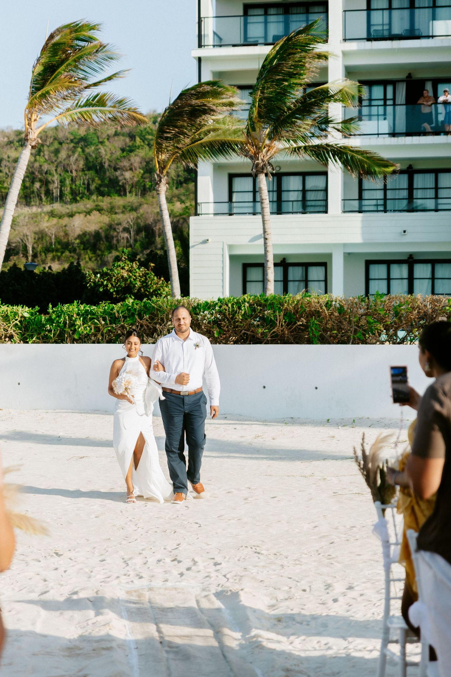 Bride and groom walk hand in hand on white sand beach with palm trees and resort building in background.