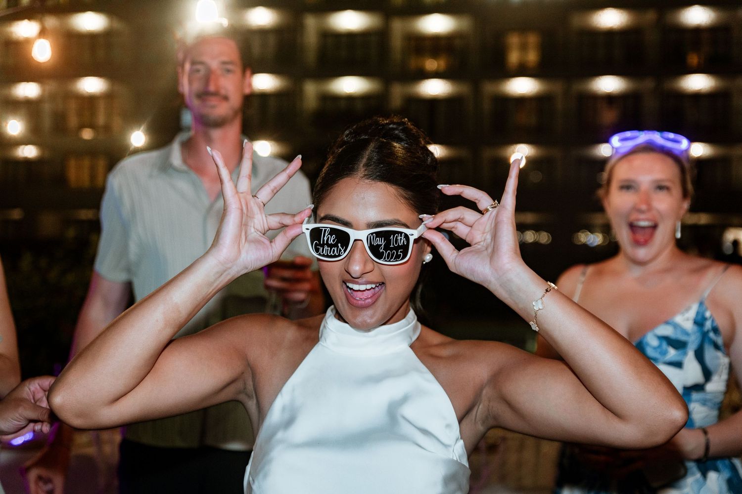Bride posing playfully with white sunglasses at lively nighttime wedding reception party.
